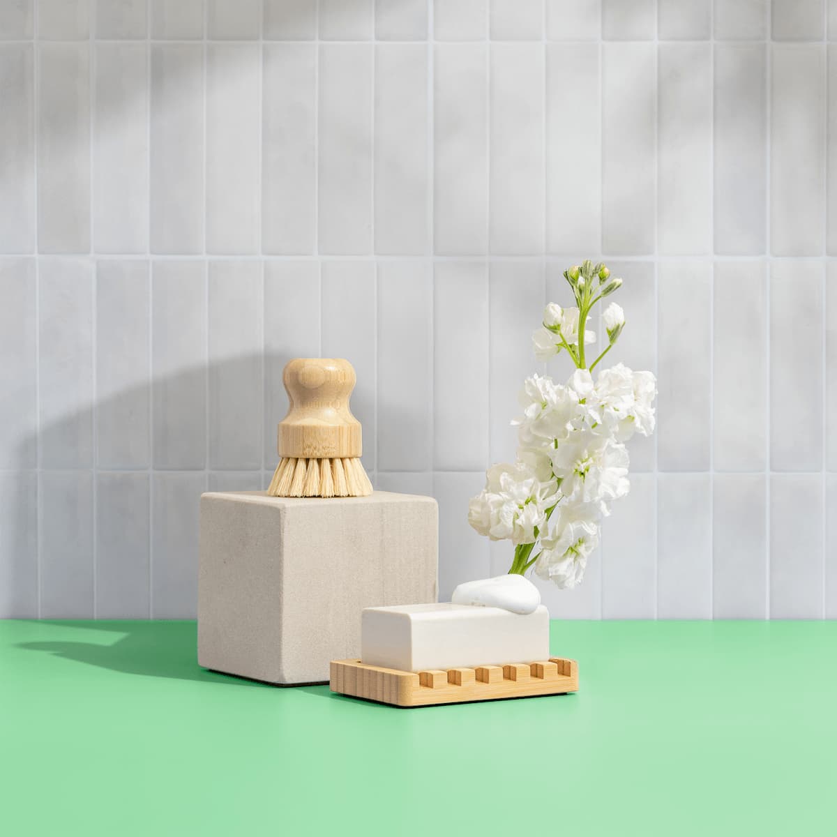 Bamboo dish brush sitting on white stone blocks and wooden soap dish, with a white dishwasher tab and white stock flowers in a vase against white subway tile on a green surface.