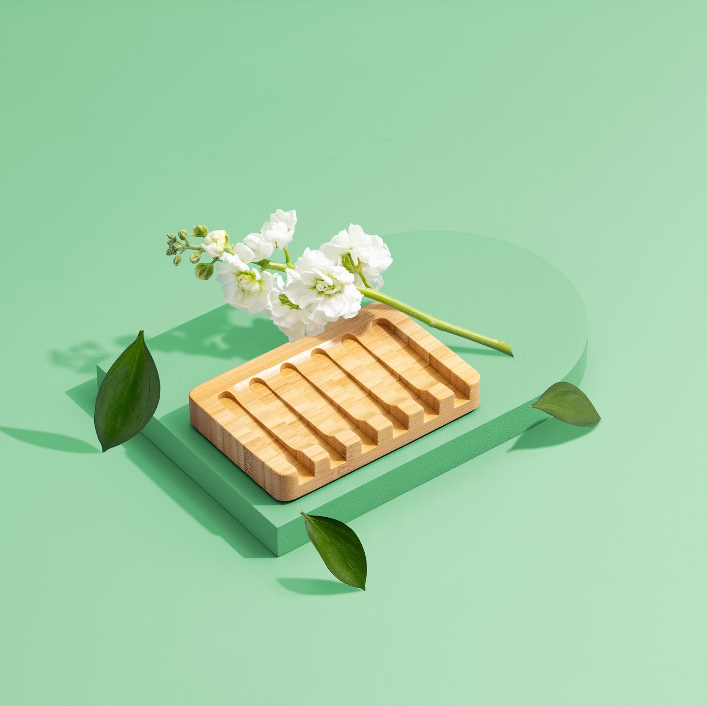 The Green Llama Bamboo Dish Soap Holder with Signature Branding rests on a green geometric platform, accented by white flowers and green leaves, set against a mint green background.