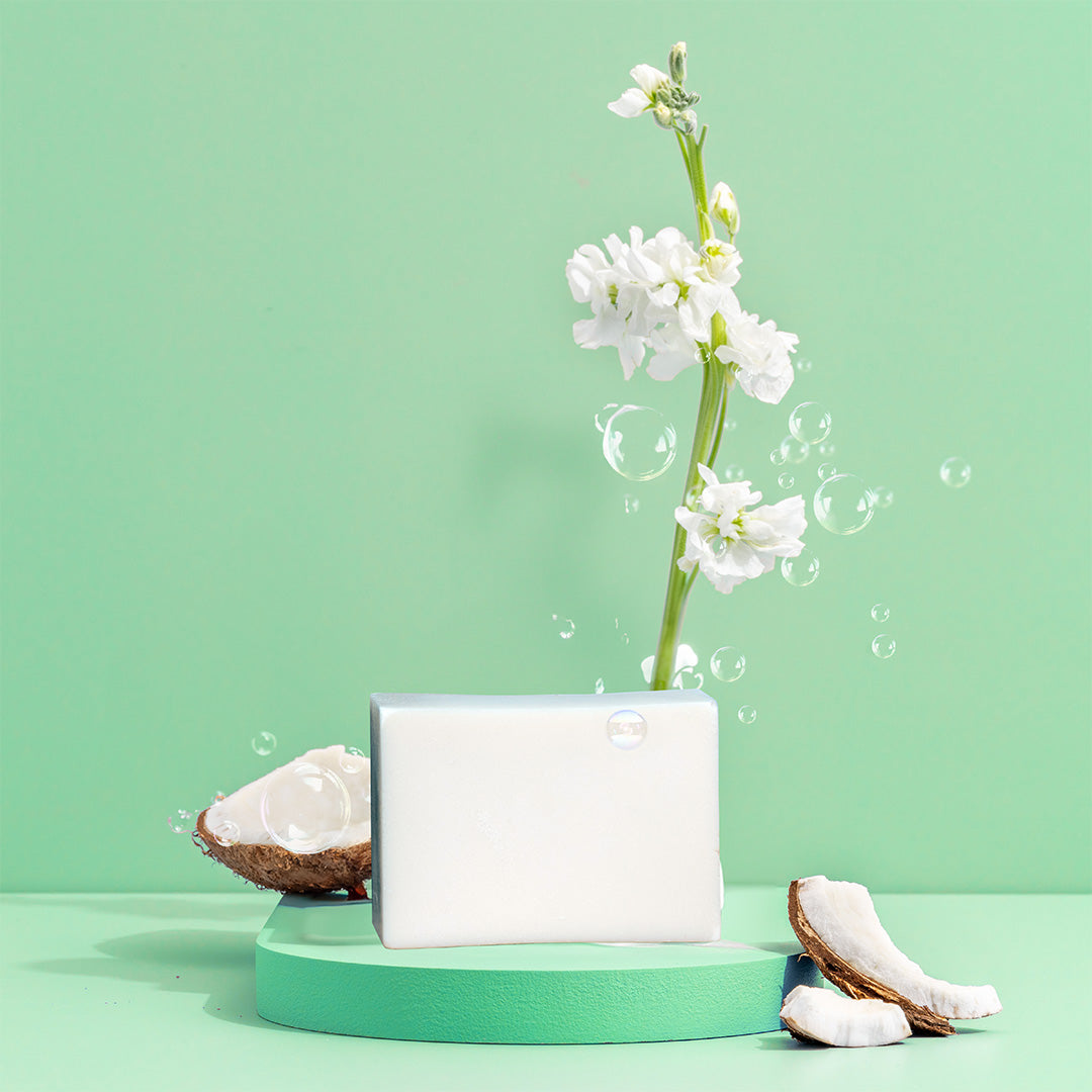 The Green Llama Solid Coconut Soap Dish Bar rests on a green display with white flowers, coconut pieces, and bubbles against a pastel green background—an eco-friendly and sustainable cleaning option.