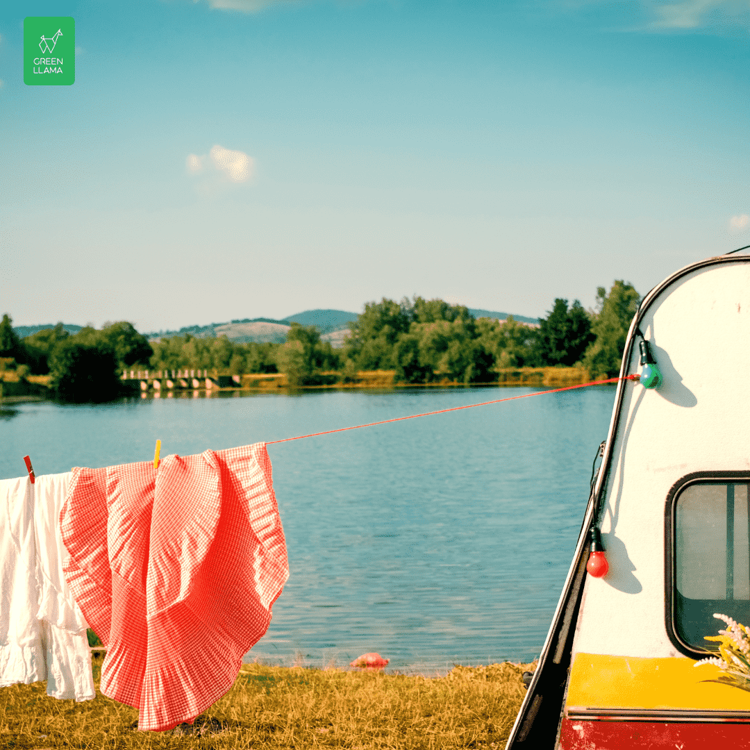 A red-and-white checked cloth and white laundry hang by a retro camper near a lake, with scenic hills behind—a perfect scene for using the Green Llama Travel Laundry & Soap Bundle by Green Llama for sustainable travel cleaning.
