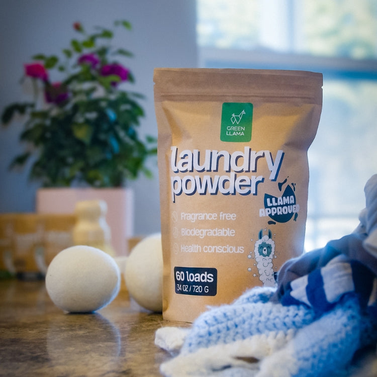 A package of Green Llama’s The Ultimate Non-Toxic Home Bulk Bundle sits on a counter with two wool dryer balls and folded laundry, highlighting plant-powered, PVA-free cleaning. A blurred potted plant and window appear in the background.