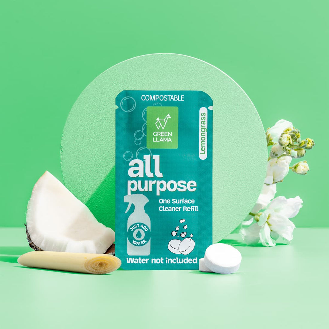 A clear spray bottle of Green Llama’s The Ultimate Home Kitchen Set stands on a green surface beside white flowers, a sliced coconut, lemongrass, and a green refill sachet—perfect for zero-waste kitchen cleaning.