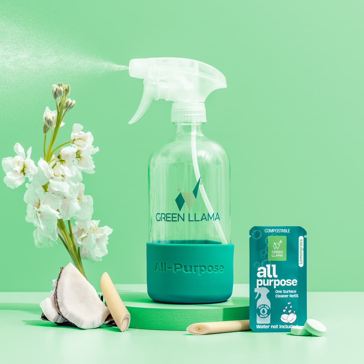 A clear spray bottle of Green Llama’s The Ultimate Home Kitchen Set stands on a green surface beside white flowers, a sliced coconut, lemongrass, and a green refill sachet—perfect for zero-waste kitchen cleaning.