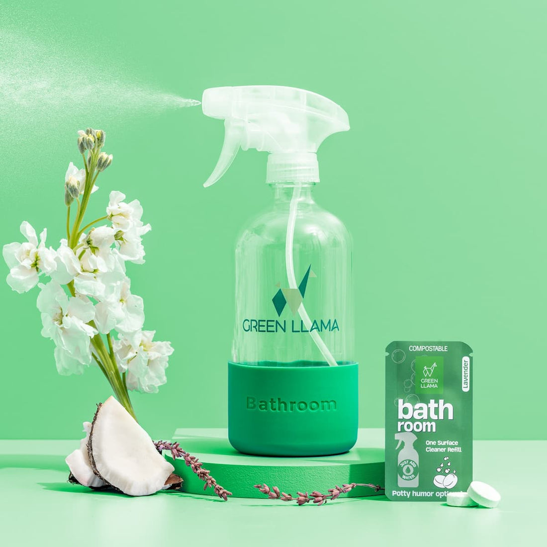 Green Llama's Bathroom Spray Glass Bottle Cleaning Kit with Lavender is shown misting near white flowers, coconut shell, and refill packet—perfect for natural bathroom cleaning—against a vibrant green background.