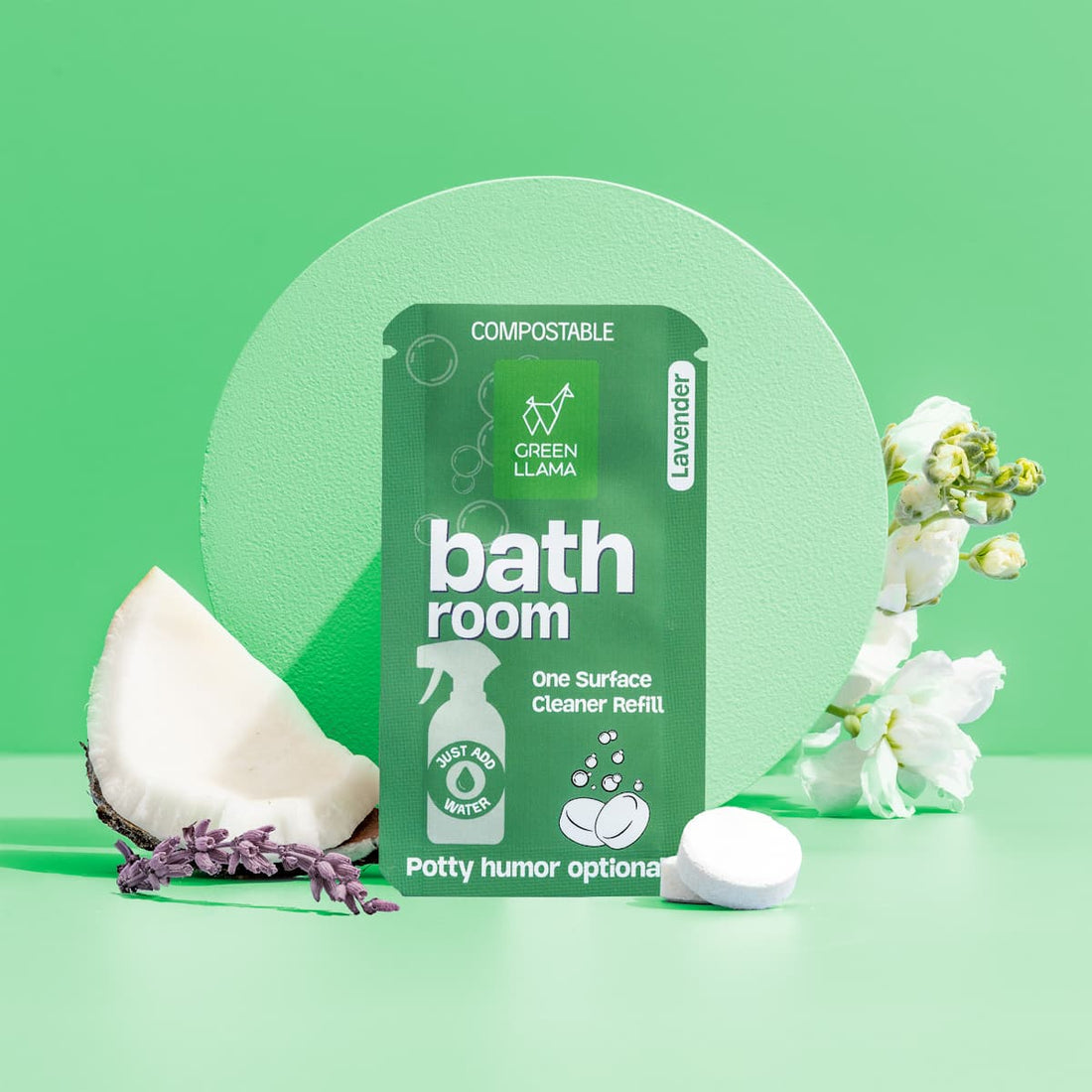 Green Llama's Bathroom Spray Glass Bottle Cleaning Kit with Lavender is shown misting near white flowers, coconut shell, and refill packet—perfect for natural bathroom cleaning—against a vibrant green background.
