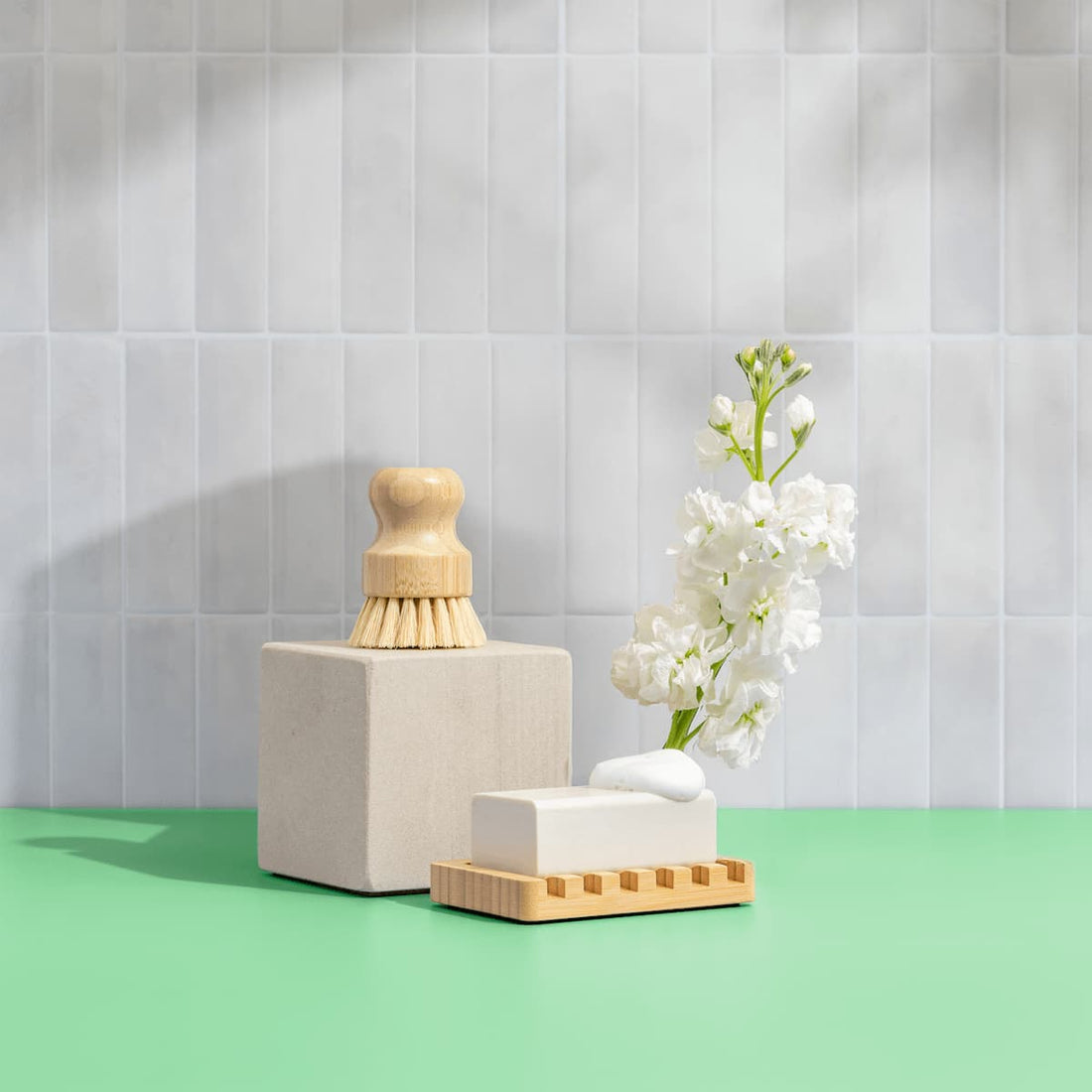 Green Llama's The Ultimate Home Kitchen Set, featuring EWG Verified soap on a wooden dish, is displayed with white flowers and a bath brush on a green surface against a light gray tiled wall.