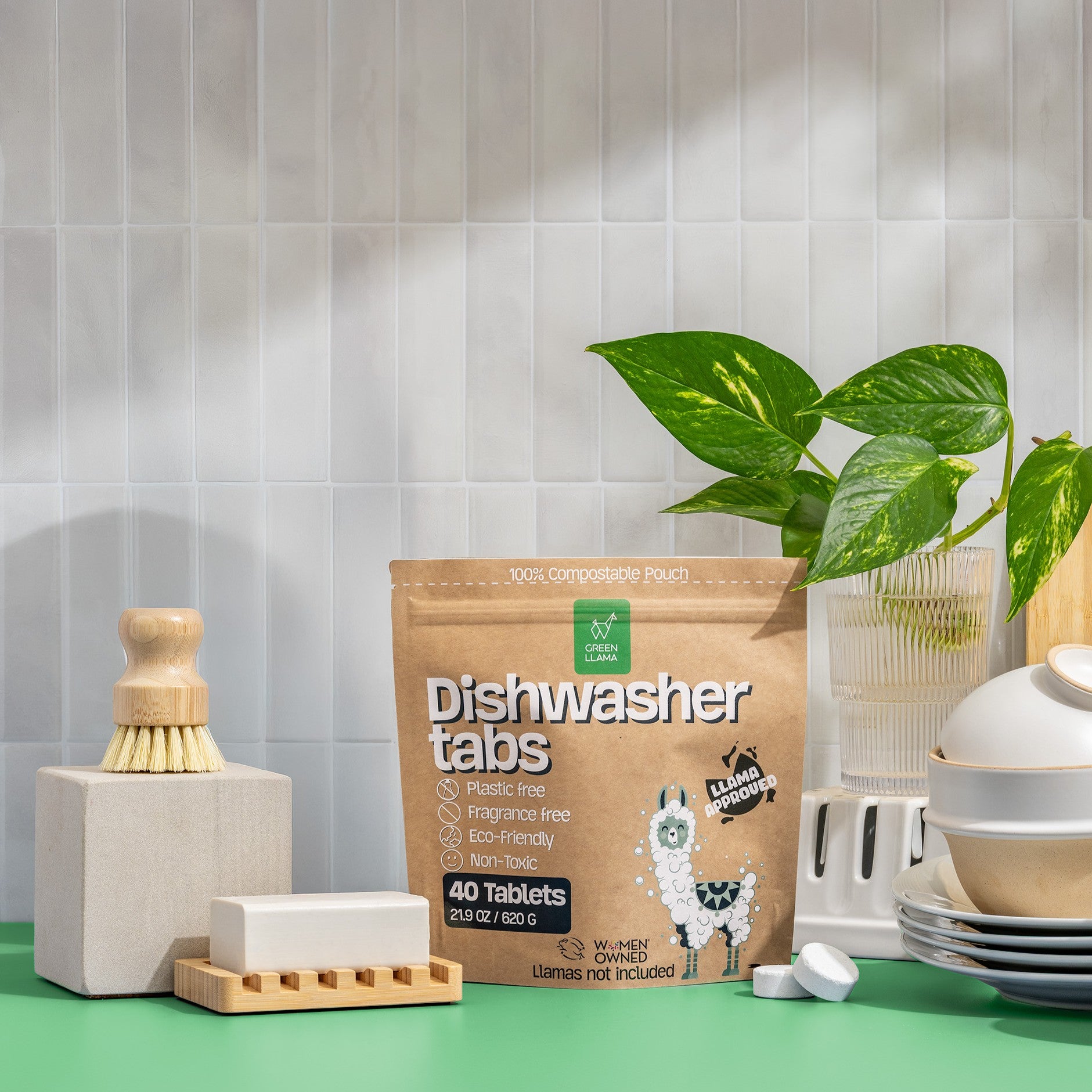 Green Llama’s Ultimate Safe & Effective Dishwashing Set sits on a counter with dishwasher tablets, a wooden dish brush, non-toxic soap in a holder, stacked plates, and a potted plant against light tiles.