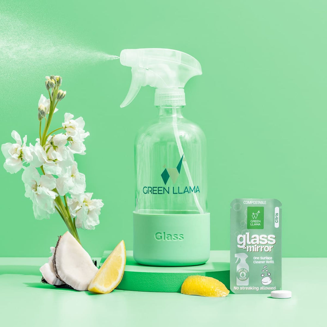 A Green Llama Starter Home Cleaner Kit—Complete 3-in-1 Eco Cleaning Kit—spray bottle sits beside a flowering branch, half coconut, lemon wedge, and refill packet, creating an eco-friendly cleaning scene on a green background.