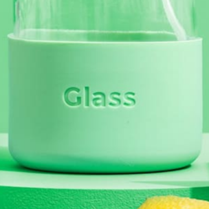 The Green Llama - Silicon Sleeve Glass Bottle Protector, featuring a light green silicone sleeve and the Green Llama logo, rests on a matching green surface with a hint of lemon nearby—a stylish and eco-friendly accessory.