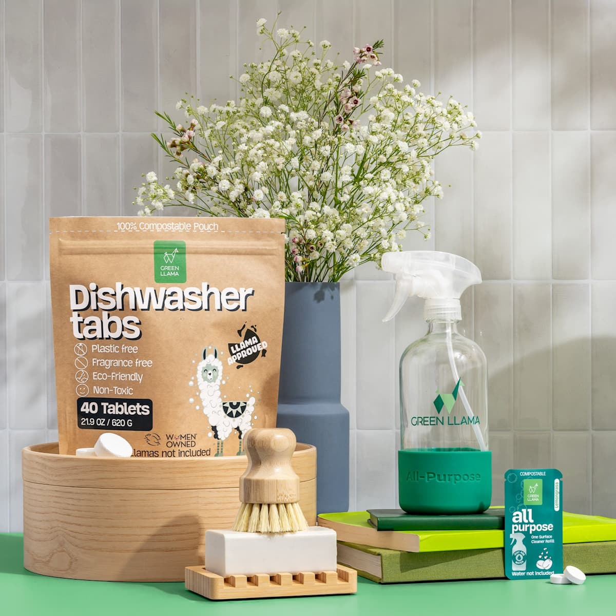 The Ultimate Home Kitchen Set by Green Llama, featuring non-toxic cleaning products for zero-waste kitchens, is displayed on a wooden tray. A vase of white flowers sits in the background.