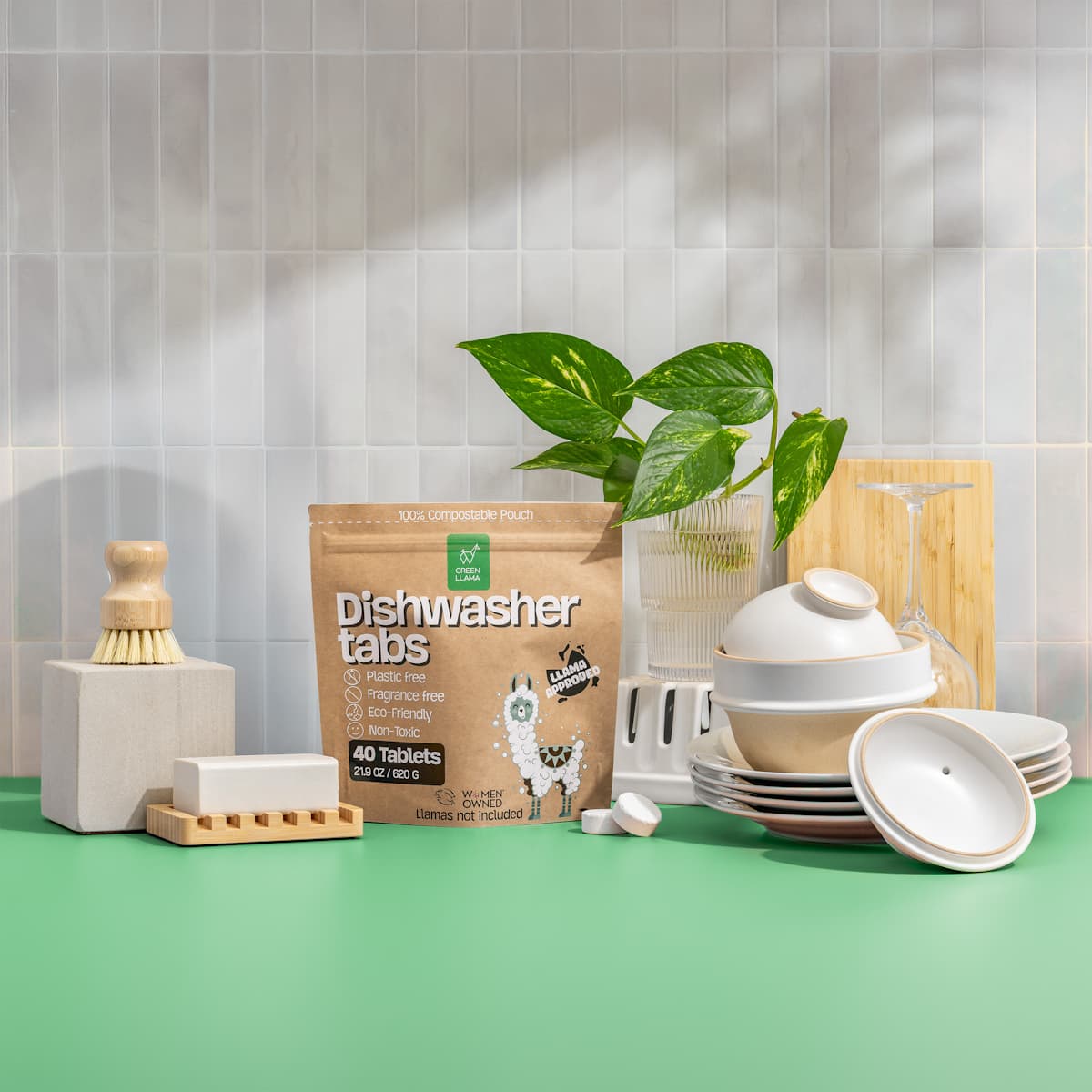Green Llama dishwasher tabs in brown compostable pouch showing '40 tablets' and eco-friendly features, arranged with stacked ceramic dishes, bamboo dish brush, wooden accessories, and a pothos plant against white subway tile on a green surface.