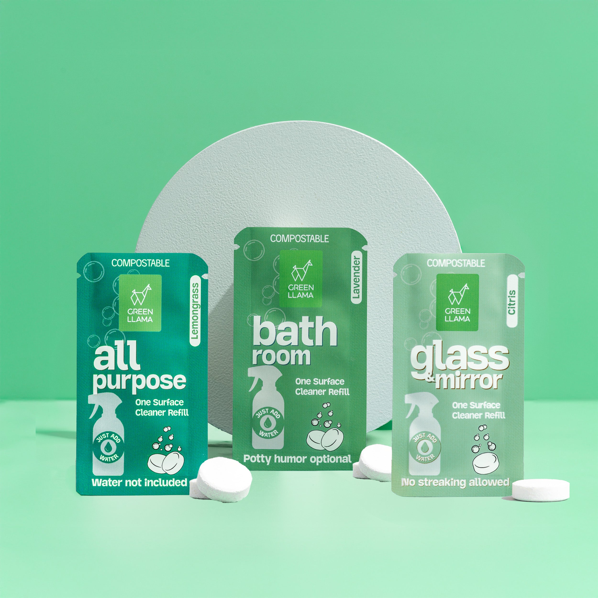 Green Llama’s Refill Cleaning Bundle features three green compostable packets—All Purpose, Bathroom, and Glass + Mirror—with eco-friendly cleaning tablets, displayed upright against a green background with a circular white backdrop.
