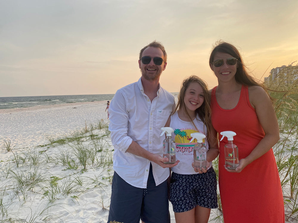 A man, a woman, and a young girl stand smiling on a sandy beach at sunset, holding glass spray bottles. The sky is clear, and sea grass is visible on the sand near the shoreline in the background.