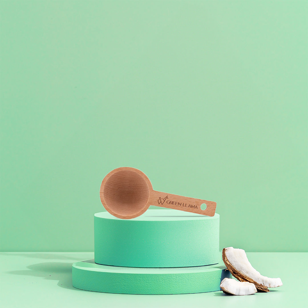 A wooden spoon engraved with Green Llama rests on mint-green cylinders. Cracked coconut pieces evoke the fresh spirit of The Complete Hypoallergenic Laundry System by Green Llama against a mint-green background.