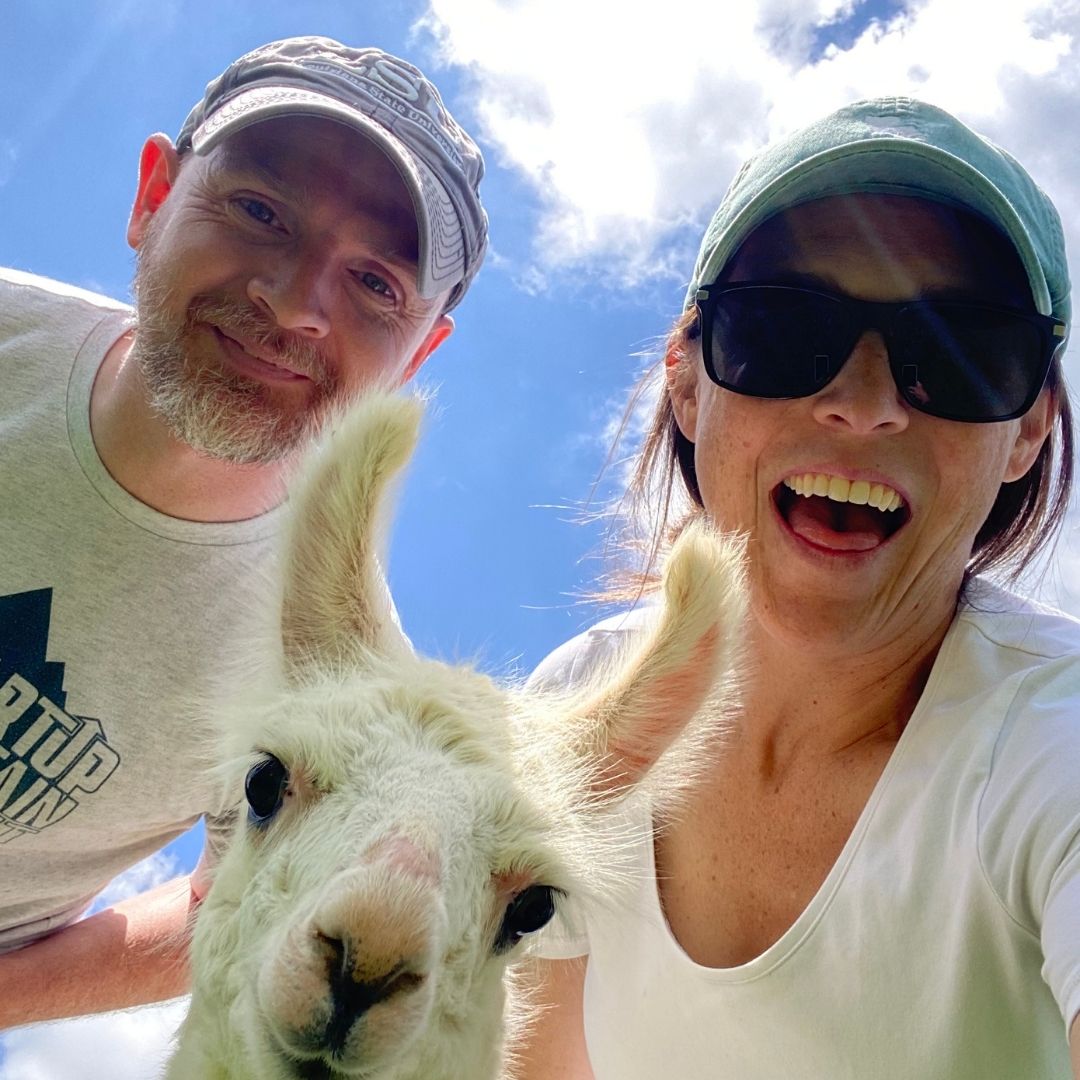 A man and a woman taking a selfie outdoors with a white alpaca in the foreground against a blue sky with clouds. The man wears a gray cap and a light gray t-shirt, and the woman wears a green cap, black sunglasses, and a white t-shirt.