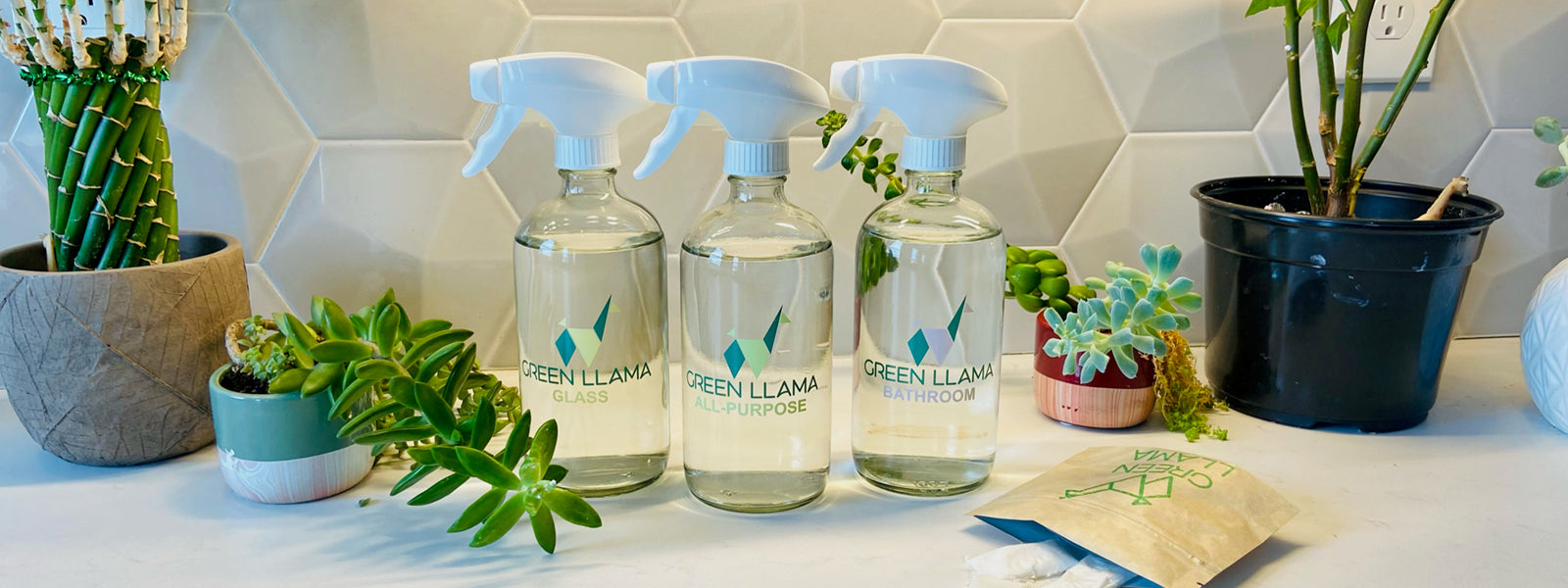 Three spray bottles labeled “Green Llama” for glass, all-purpose, and bathroom cleaning sit on a white counter with potted plants and succulents, in front of a hexagonal tile backsplash.