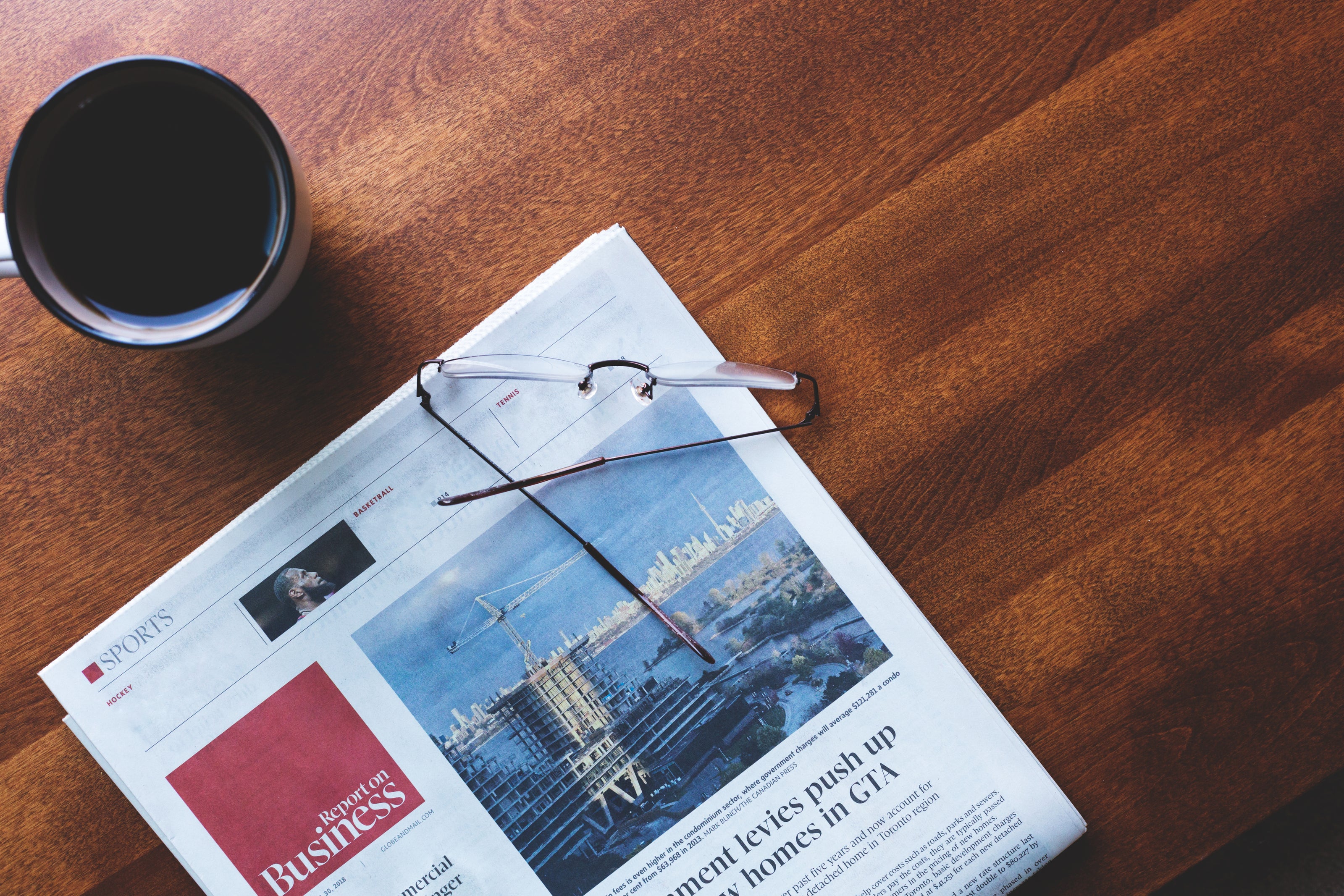 A pair of eyeglasses rests on a folded newspaper featuring business news and a cityscape photo, next to a cup of coffee on a wooden table.