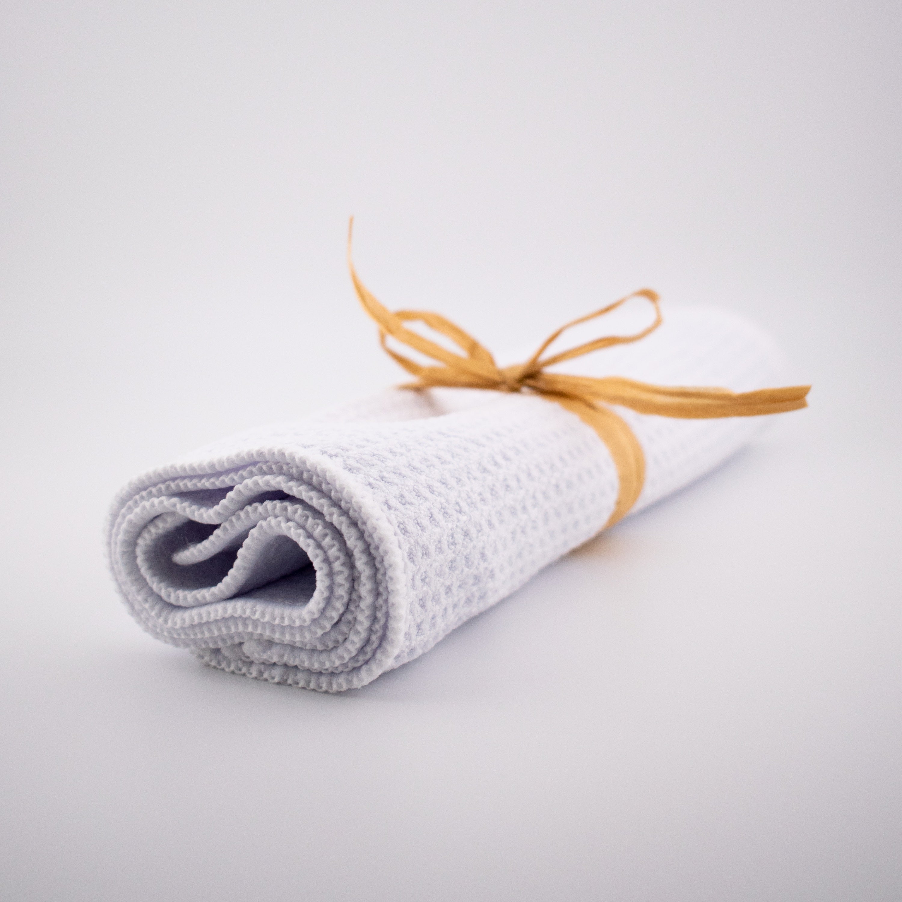 A neatly rolled Green Llama High-Performance Organic Cotton Cleaning Towel (from the 3-Pack), white with a waffle texture and tied with a light brown raffia ribbon, sits on a plain white background.