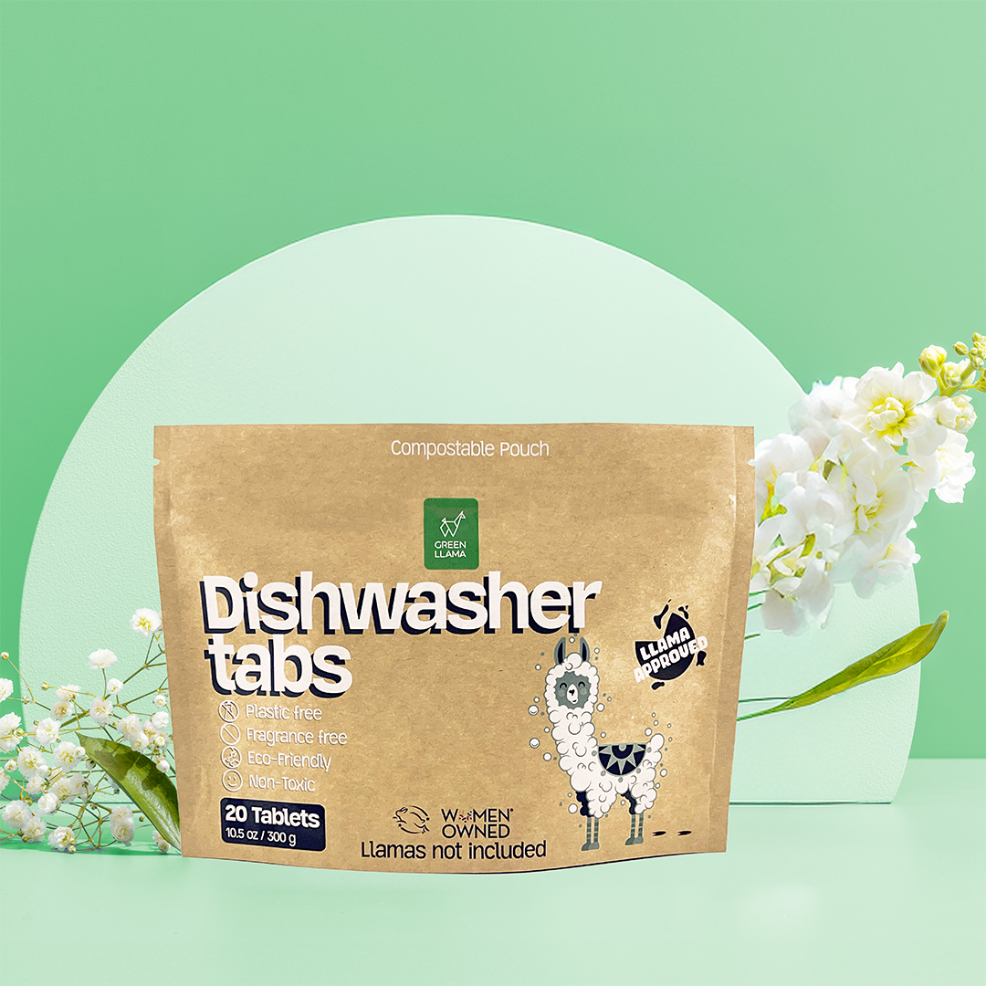 A compostable pouch labeled “EWG Verified™ & PVA-Free Dishwasher Tabs” by Green Llama stands upright on a green surface with white flowers nearby. The plastic-free packaging features a cartoon llama with suds, showcasing its eco-friendly appeal.