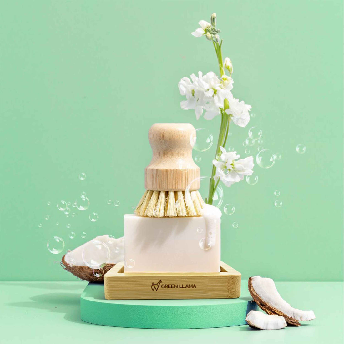Green Llama's The Ultimate Home Kitchen Set, featuring EWG Verified soap on a wooden dish, is displayed with white flowers and a bath brush on a green surface against a light gray tiled wall.