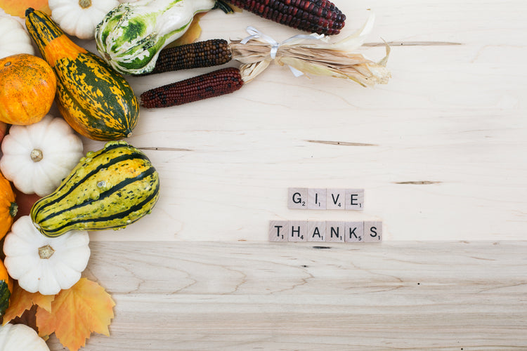 Assorted gourds, miniature pumpkins, and colorful corn arranged on a wooden surface with the phrase GIVE THANKS spelled out in Scrabble tiles on the right side.