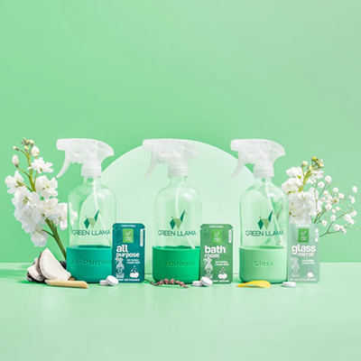Three Green Llama spray bottles labeled All-Purpose, Bathroom, and Glass are displayed with matching cleaner refill packets in front, surrounded by white flowers and a green background.