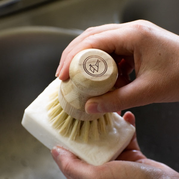 A person uses the Green Llama Valentine's Special Complete Dish Washing Starter Bundle—Solid Soap, Bamboo Holder, & Scrub Brush—scrubbing the eco-friendly bar made with Saponified Coconut Oil. The wooden brush handle features an engraved logo.