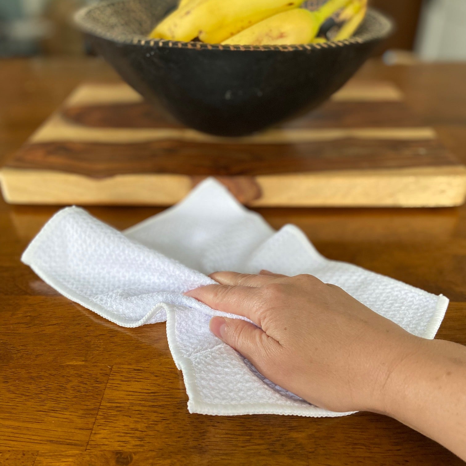A hand uses a Green Llama High-Performance Organic Cotton Cleaning Towel (3-Pack) to wipe a wooden table, with a bowl of yellow bananas on a cutting board in the background.